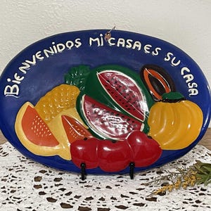 May include: A blue ceramic plate with a white border and the text "Bienvenidos Mi Casa Es Su Casa" in white. The plate is decorated with a colorful fruit design, including watermelon, pineapple, and bananas.
