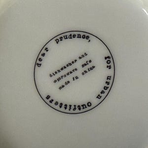 May include: A white ceramic bowl with a black circular stamp. The stamp reads "dear prudence, dishwasher and microwave safe, made in china, for urban outfitters."