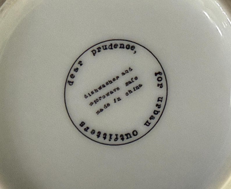 May include: A white ceramic bowl with a black circular stamp. The stamp reads "dear prudence, dishwasher and microwave safe, made in china, for urban outfitters."