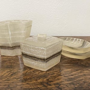 May include: A set of three bathroom accessories made of clear, translucent, striped, and textured plastic. The set includes a toothbrush holder, a lidded jar, and a soap dish.