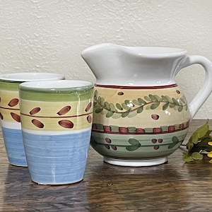 May include: A white pitcher with a green, yellow, and red floral design. Two matching mugs with the same design are next to the pitcher.