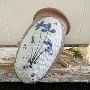 May include: A white ceramic vase with a brown rim and blue floral design. The vase has a textured surface and is slightly tilted.