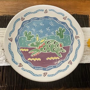 May include: A white ceramic plate with a colorful design of a green rabbit with a purple, yellow, and blue background. The plate has a blue and white wavy border with a geometric pattern. The plate is signed "Geiger" in the bottom right corner.