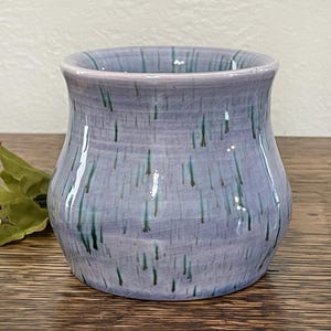 May include: A small, light purple ceramic vase with a textured, speckled glaze. The vase has a wide base and a narrow opening, and it is decorated with thin, green lines.