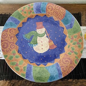 May include: A round ceramic plate featuring a central dark blue star-shaped medallion with a white snowman wearing a red top hat and a green scarf, holding a broom. The border displays a colorful patchwork pattern of swirls, holly, and stripes.