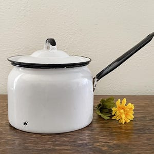 May include: White enamel saucepan with a black handle and black rim. The saucepan has a lid with a black knob.