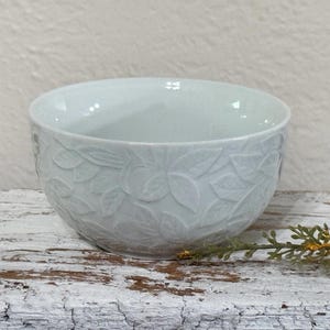 May include: A light blue ceramic bowl with a detailed embossed floral pattern. The bowl has a smooth interior and a textured exterior with a repeating leaf design. The bowl is sitting on a distressed white wooden surface.
