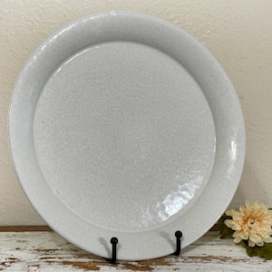 May include: A round, light gray ceramic plate with a textured surface. The plate has a raised, circular center and a wide, flat rim. The plate is displayed on a black metal stand, with a decorative flower in the foreground. The plate is suitable for serving food.
