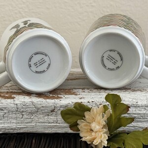 May include: Two white ceramic mugs with handles, resting on their rims. One mug features a design of birds and foliage, the other has a geometric pattern. The bottoms display text within a circular border.