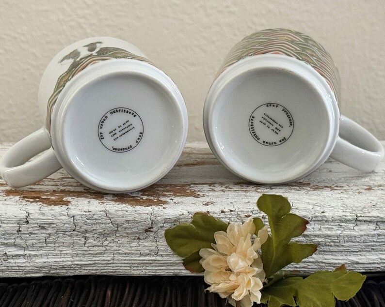 May include: Two white ceramic mugs with handles, resting on their rims. One mug features a design of birds and foliage, the other has a geometric pattern. The bottoms display text within a circular border.