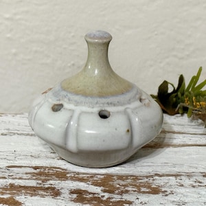 May include: A ceramic incense burner with a bulbous, white body and a light brown top. The burner has three small holes and a small, rounded handle. The incense burner is on a distressed, white wooden surface.