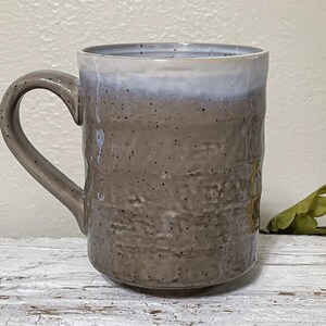 May include: A speckled gray ceramic mug with a white rim and a handle. The mug has a textured surface and a slightly faded white glaze.