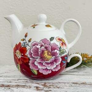 May include: White ceramic teapot with a matching teacup. The teapot and teacup are decorated with a large pink and red floral design. The teapot has a lid and a handle. The teacup is attached to the teapot.