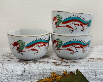 May include: Three white ceramic teacups with gold rims and a red, blue, and green dragon design.