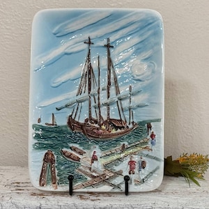 May include: A rectangular ceramic wall plaque featuring a raised relief scene of a brown two-masted sailing ship on textured green water. The background shows a light blue sky with white clouds, while the foreground depicts a wooden dock.