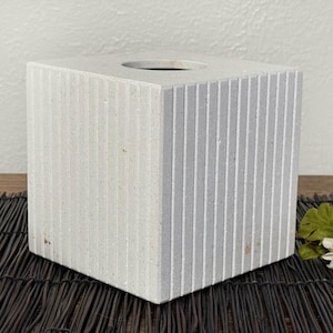 May include: A square, light gray tissue box cover with vertical, grooved lines. The top has a circular opening for tissue dispensing. The box is made of a stone-like material and sits on a dark brown surface.