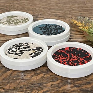 May include: Four round white coasters with different patterned fabric inserts. The coasters have a black and white floral pattern, a black and teal floral pattern, a black and red floral pattern, and a beige and black floral pattern.
