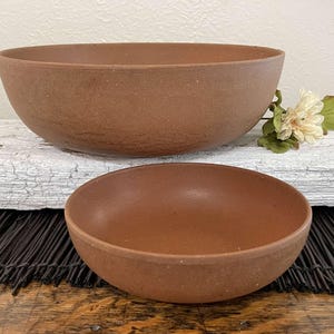 May include: Two brown ceramic bowls of different sizes. The larger bowl is in the background, and the smaller bowl is in the foreground. The bowls have a textured surface and a simple, rounded design. A flower with white petals and green leaves is in the background.