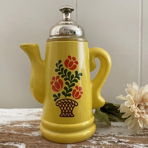 May include: Yellow ceramic teapot with a silver lid and floral design. The teapot has a spout and a handle. The flowers are red, green, and yellow.