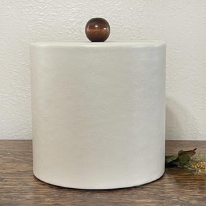 May include: A cylindrical, off-white container with a textured surface and a dark brown wooden knob on top. The container is sitting on a wooden surface.
