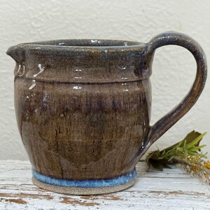 May include: A ceramic creamer pitcher featuring a glossy brown glaze with subtle purple and blue undertones. The pitcher has a rounded body, a small pouring spout, a curved handle, and a distinct bright blue band around the base.