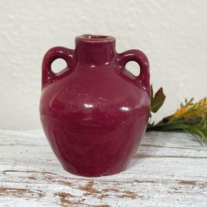 May include: A small, deep burgundy ceramic pitcher with two handles. The pitcher has a slightly rounded body and a smooth, glossy finish.