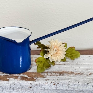 May include: A cobalt blue enamelware coffee pot with a long handle and white interior. The pot is displayed with a cream-colored flower and green leaves on a distressed, white-painted wooden surface. A vintage kitchenware item.
