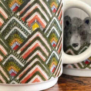 May include: White ceramic mug with a colorful geometric pattern in shades of green, orange, yellow, and white. The mug features a detailed illustration of a bear. The handle is white.