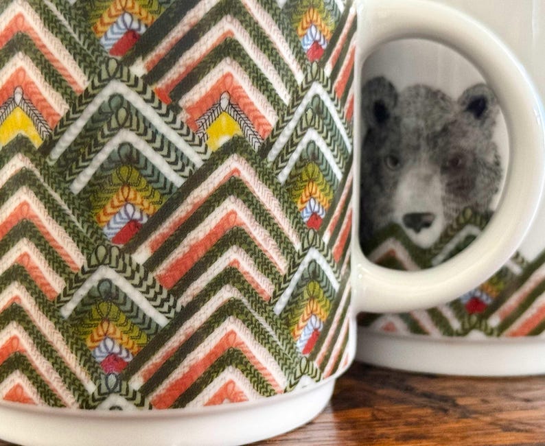 May include: White ceramic mug with a colorful geometric pattern in shades of green, orange, yellow, and white. The mug features a detailed illustration of a bear. The handle is white.