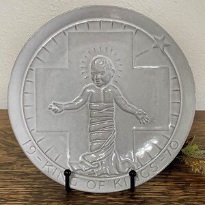 May include: A white ceramic plate with a raised image of a baby Jesus on a cross. The plate is decorated with a star and the words "King of Kings 1970".