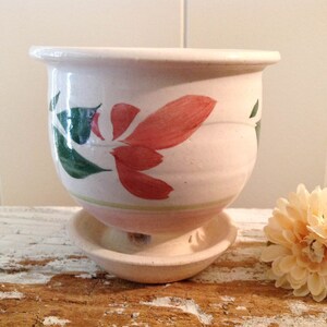 May include: A ceramic planter with a matching saucer. The off-white planter features hand-painted red and green floral designs. The planter has a rounded shape with a slightly flared rim. The saucer is a simple, shallow dish.