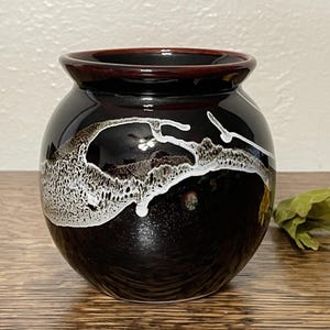 May include: A small, round, black ceramic vase with a white glaze design. The vase has a dark red rim.