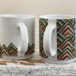May include: Two white ceramic mugs with a geometric pattern in green, orange, and yellow. The mugs have a curved handle and a clean, modern design. The pattern is located at the bottom of the mugs.