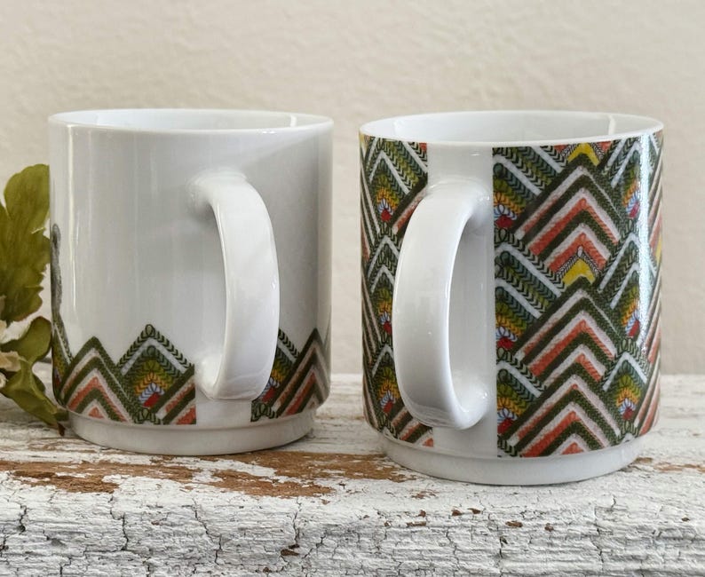 May include: Two white ceramic mugs with a geometric pattern in green, orange, and yellow. The mugs have a curved handle and a clean, modern design. The pattern is located at the bottom of the mugs.