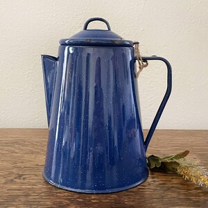 May include: A blue enamelware coffee pot with a lid and handle. The pot has a speckled finish and a spout for pouring. The handle is attached with a natural fiber. The pot is sitting on a wooden surface.