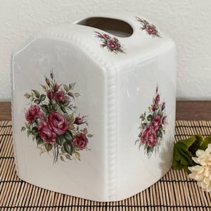 May include: A white ceramic tissue box cover with a rounded top and a central oval opening. The exterior features a floral pattern of pink roses with green leaves on the front and top panels, accented by a subtle embossed rope-like border detail.