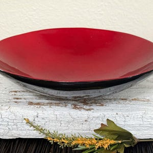 May include: A decorative, shallow bowl with a glossy red interior and a black rim. The bowl has a smooth, curved design and sits on a white, textured surface. The bowl is empty and ready to be filled.