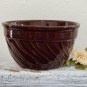 May include: A dark brown ceramic mixing bowl with a spiral design. The bowl has a wide rim and a textured exterior. A cream-colored flower with green leaves rests on the surface next to the bowl.