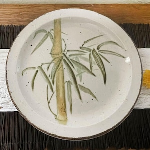 May include: A round, off-white ceramic plate with a hand-painted bamboo design. The bamboo stalk is tan, and the leaves are green. The rim of the plate has a brown speckled edge. The plate is on a dark brown mat.