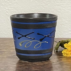 May include: A small, dark blue ceramic cup with black and white abstract designs. The cup has a wide rim and a rounded bottom.