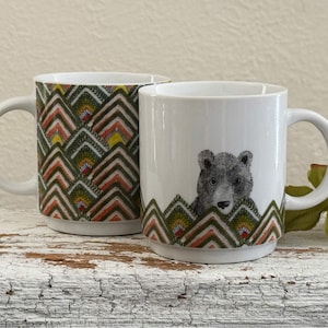 May include: Two white ceramic mugs. One mug features a repeating geometric pattern in shades of green, yellow, orange, and brown. The other mug has a gray bear illustration and a matching geometric pattern at the base.