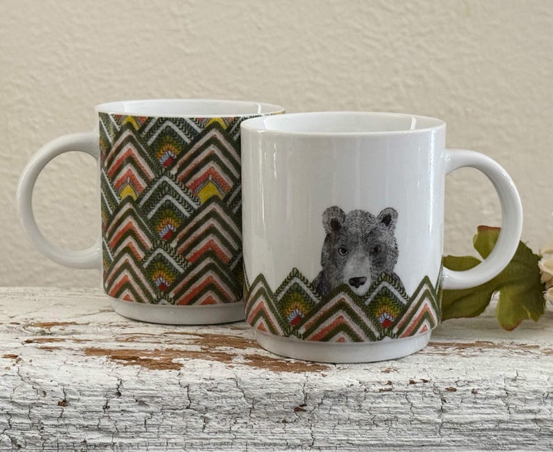 May include: Two white ceramic mugs. One mug features a repeating geometric pattern in shades of green, yellow, orange, and brown. The other mug has a gray bear illustration and a matching geometric pattern at the base.
