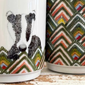 May include: Two white ceramic mugs. One mug features a badger illustration in black and white. The other mug has a colorful geometric pattern in green, orange, yellow, and white. Both mugs have a decorative chevron design at the base.