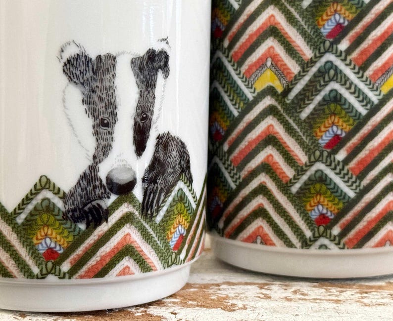 May include: Two white ceramic mugs. One mug features a badger illustration in black and white. The other mug has a colorful geometric pattern in green, orange, yellow, and white. Both mugs have a decorative chevron design at the base.