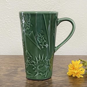 May include: A tall green ceramic mug with a handle. The mug has a textured, abstract design with a raised pattern.