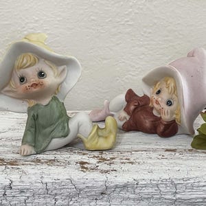 May include: Two ceramic elf figurines. One elf wears a green shirt, white pants, and a yellow hat and shoes. The other elf wears a brown shirt, white pants, and a pink hat. Both have blonde hair and are lying down.