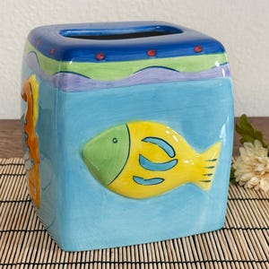 May include: A square ceramic tissue box featuring a colorful aquatic design. The box is painted light blue with a yellow fish and a seahorse. The top has a blue, green, and purple wavy design. The tissue box is a decorative home decor item.