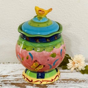 May include: A colorful ceramic jar with a lid, featuring a yellow bird on top. The jar is painted with pink, blue, green, and yellow accents, and a butterfly design. The jar sits on a decorative base.