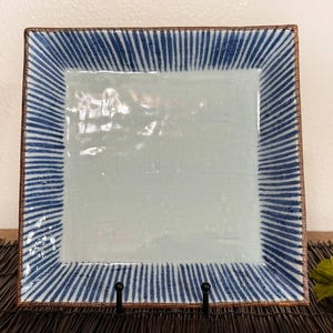 May include: A square ceramic plate featuring a smooth, light blue glazed center surrounded by a wide border decorated with thin, vertical dark blue stripes. The edges of the plate have a distinct brown, textured finish along the outer rim.