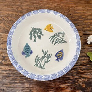 May include: A white ceramic pie plate with a blue and white border. The plate features a hand-painted design of colorful fish and coral.